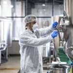 Technologist in protective uniform wearing hairnet mask goggles and gloves closing industrial tank reservoir.